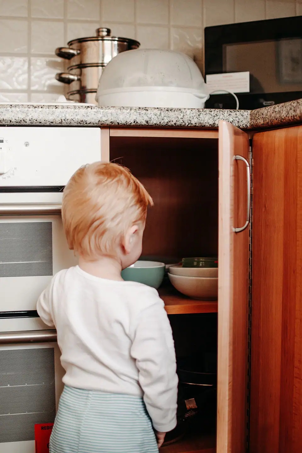 A toddler, seen from behind, looking into an open low kitchen cabinet full of bowls